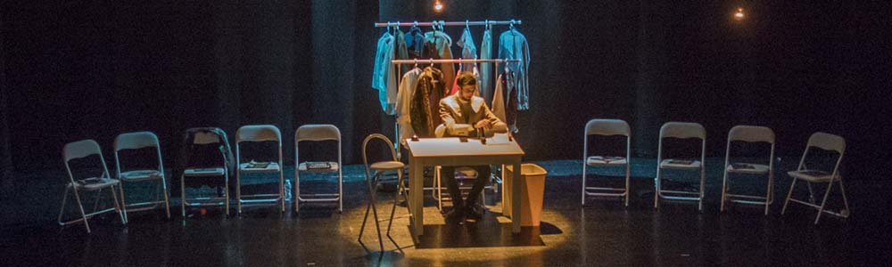 Un home sentado con diversos elelemntos ao seu redor nunha representación teatral